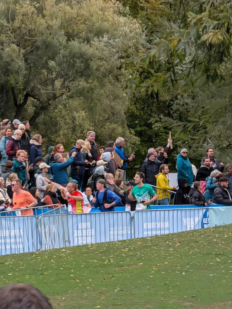Spectators lining the Munich Marathon 2026 course in the Olympiapark as Oly Bock runs past in a green shirt