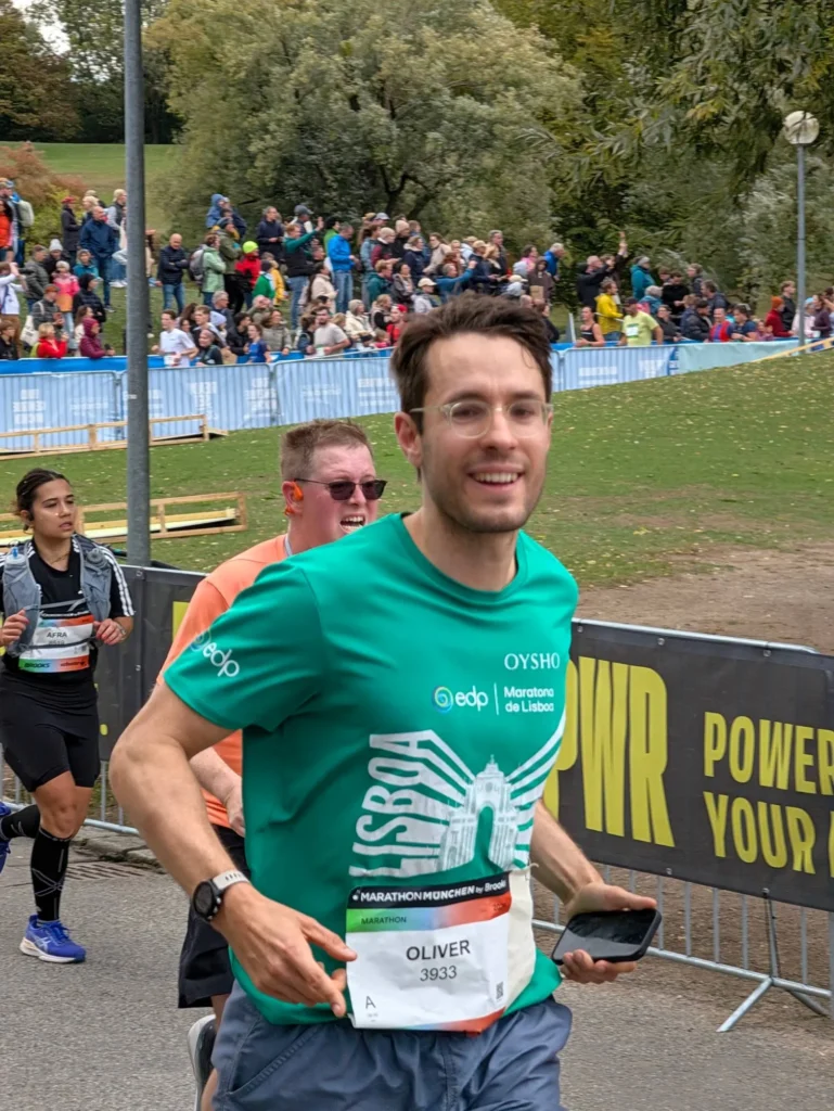 Oly Bock smiling while running the Munich Marathon 2026 in the Olympiapark, wearing a green Lisbon Marathon shirt and bib 3933
