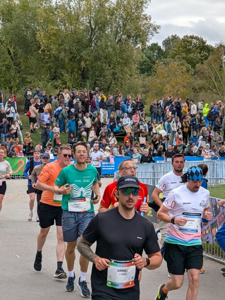 Oly Bock in a green Lisbon Marathon shirt among other runners at the Munich Marathon 2026, spectators cheering on the hill behind